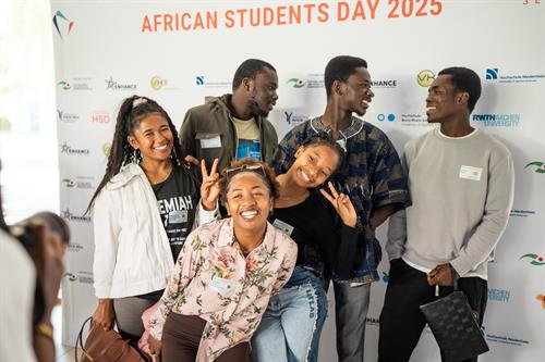 Sechs junge Erwachsene posieren lachend vor einer Fotowand mit der Aufschrift „African Students Day 2025“. Zwei Personen im Vordergrund machen ein Peace-Zeichen, während andere sich zu ihnen umdrehen und miteinander interagieren. Die Stimmung ist freundlich und ausgelassen.